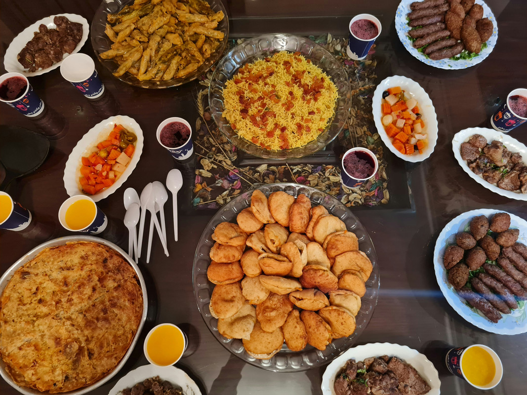 An iftar food selection inluding pickles, kebbeh, kebabs, fried samosas, juices and plastic spooins.